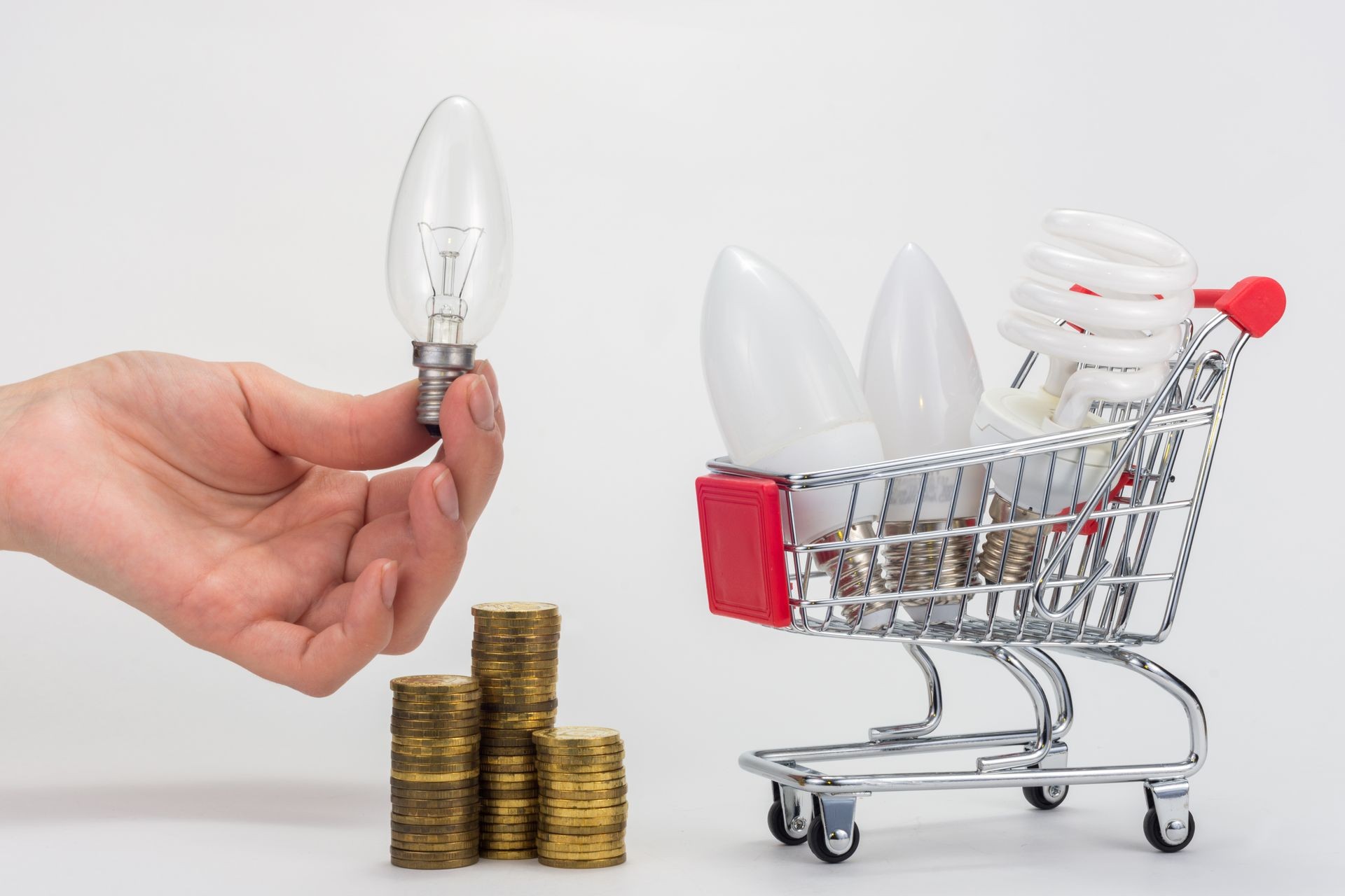In the grocery cart are LED and energy-saving light bulbs, next to a hand is holding an incandescent lamp, there is a stack of coins